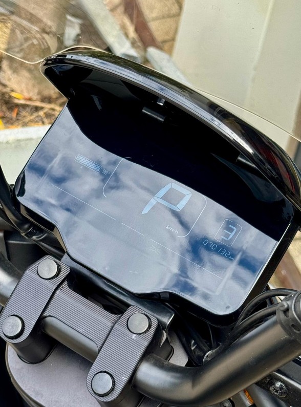 Digitaal dashboard van een motorfiets met P-indicator, batterij-icoon en stuurklem, met wolken weerspiegeld.