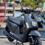 Zwarte Tomos-scooter op de standaard, geparkeerd op de stoep voor een winkelraam.