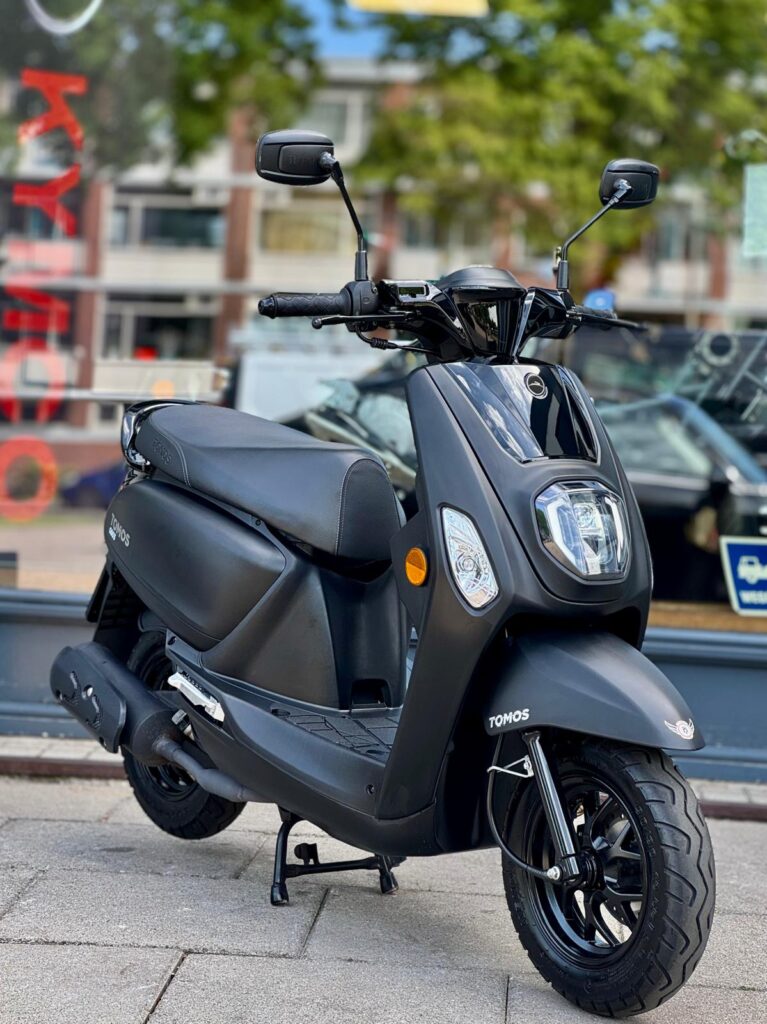 Zwarte Tomos-scooter op de standaard, geparkeerd op de stoep voor een winkelraam.