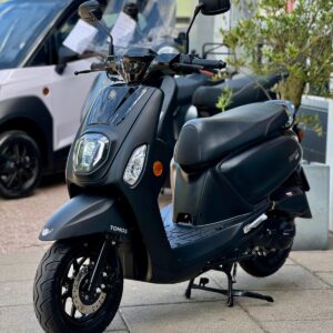 Zwarte Tomos-scooter op de stoep voor een showroom, met schijfrem voor en de spiegels omhoog.