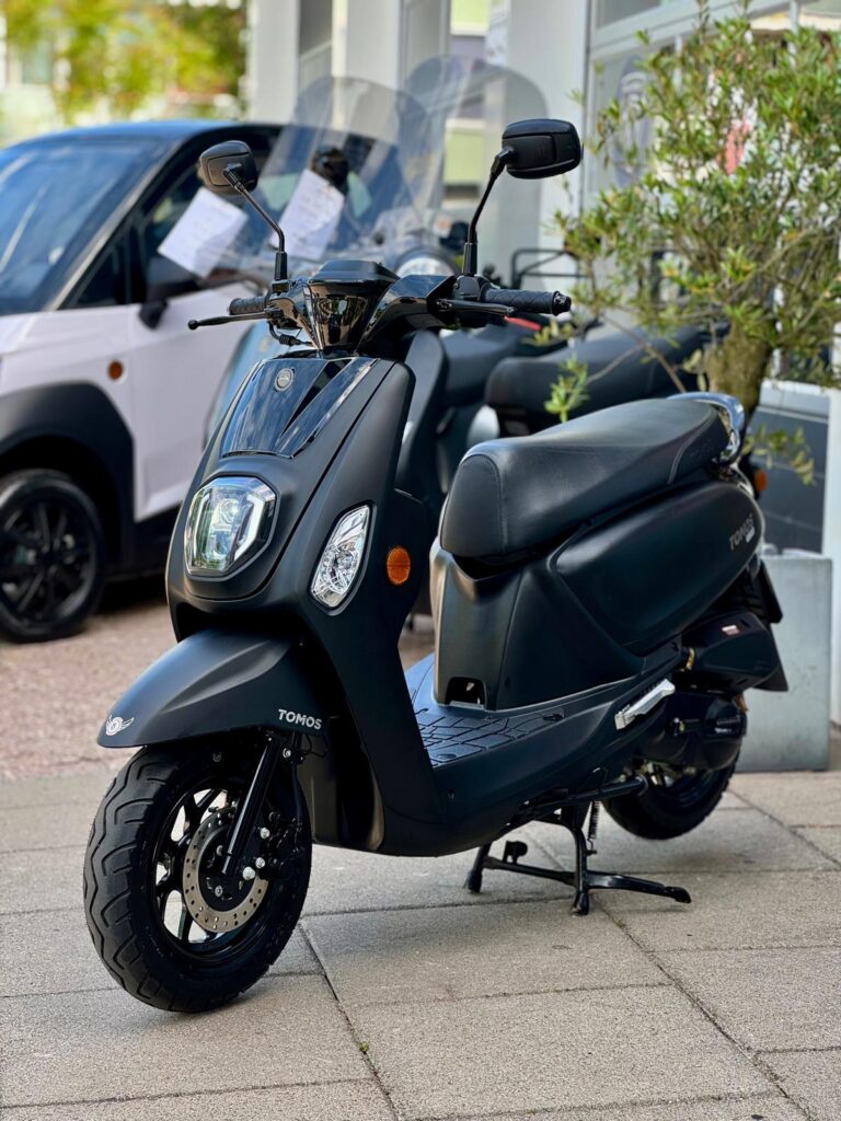 Zwarte Tomos-scooter op de stoep voor een showroom, met schijfrem voor en de spiegels omhoog.