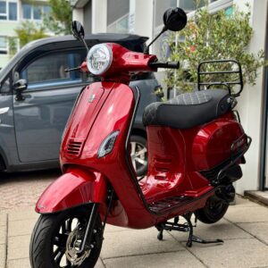 Rode retro-scooter geparkeerd op stoep voor garage, met grijze auto op achtergrond.