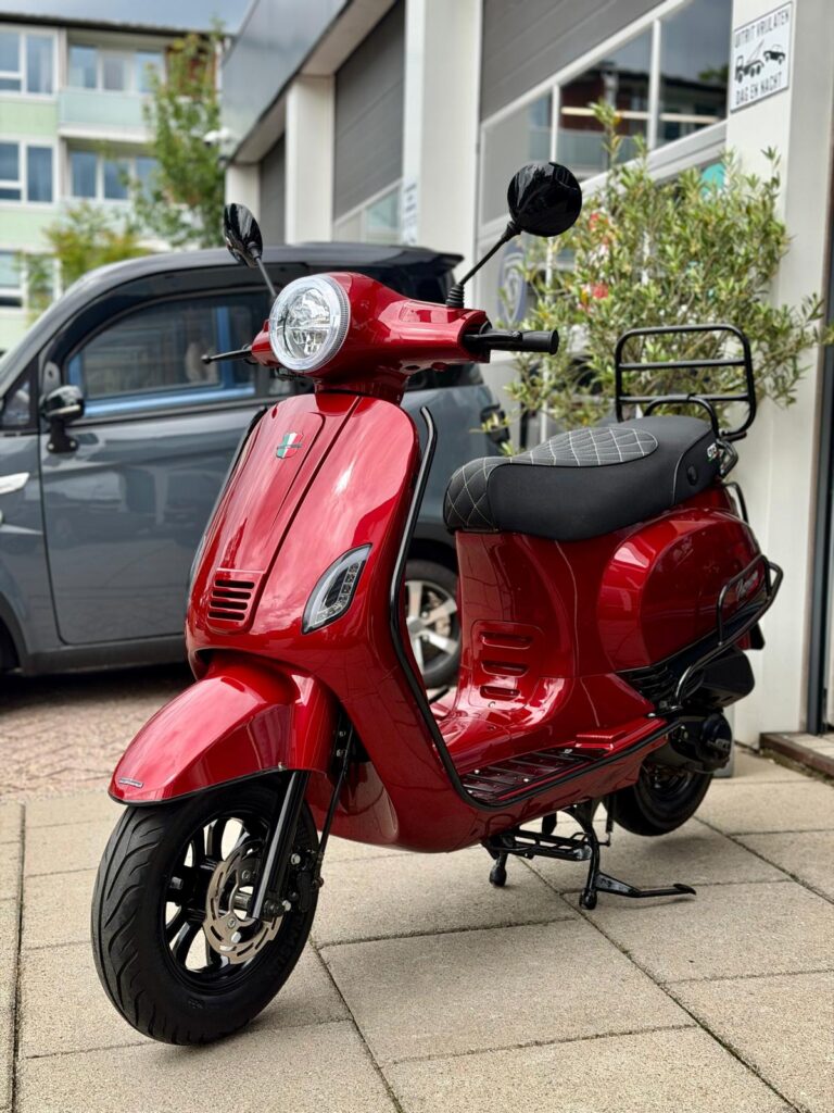 Rode retro-scooter geparkeerd op stoep voor garage, met grijze auto op achtergrond.