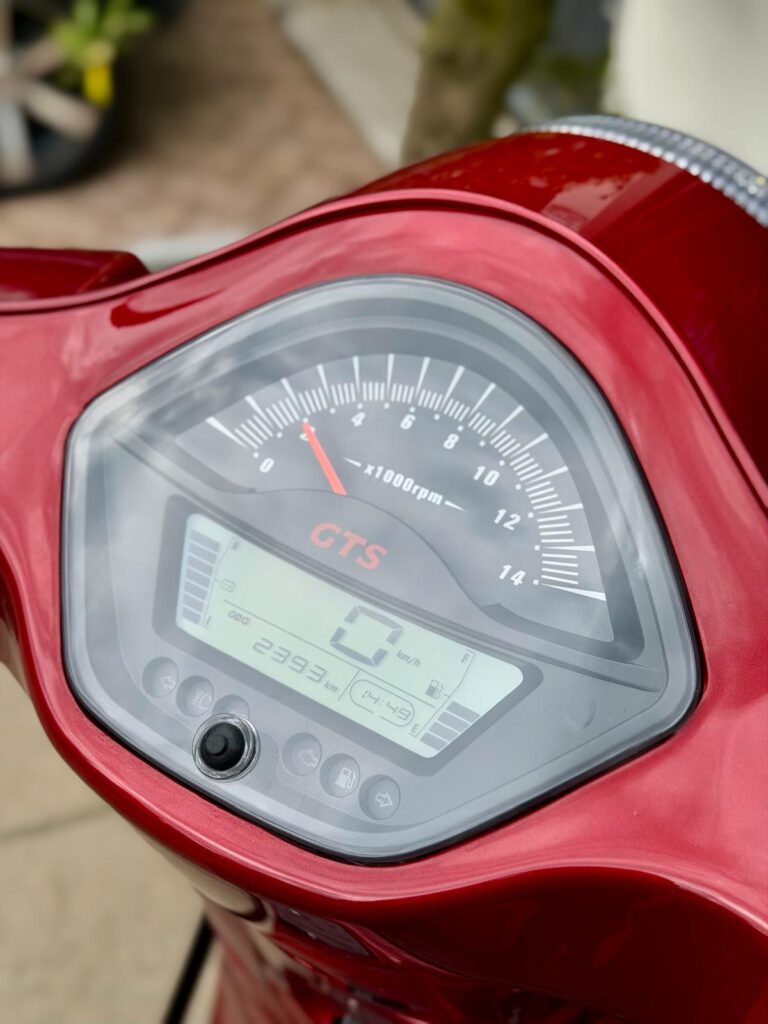 Close-up van het dashboard van een rode scooter met analoge toerenteller en digitaal scherm met 0 km/u.