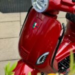 Close-up van een glanzende rode scooter met ronde koplamp, Italiaans schildembleem en planten op de voorgrond.