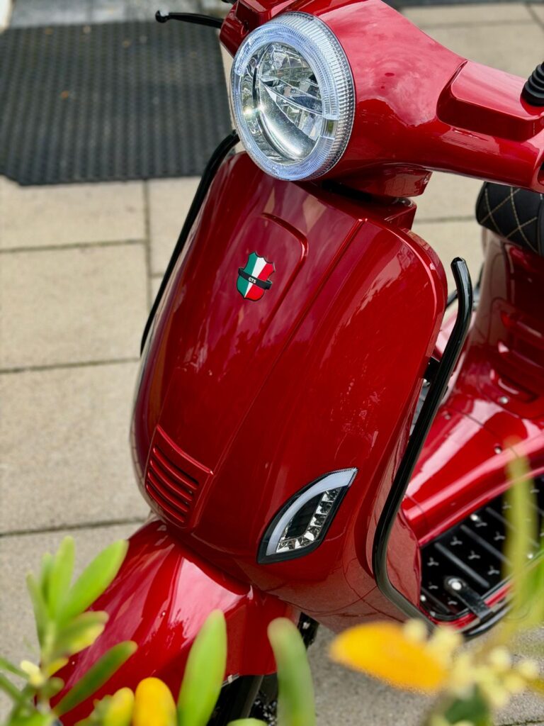 Close-up van een glanzende rode scooter met ronde koplamp, Italiaans schildembleem en planten op de voorgrond.