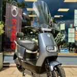 Moderne grijze scooter met windscherm, geparkeerd voor een motorzaak met Kymco-banner.