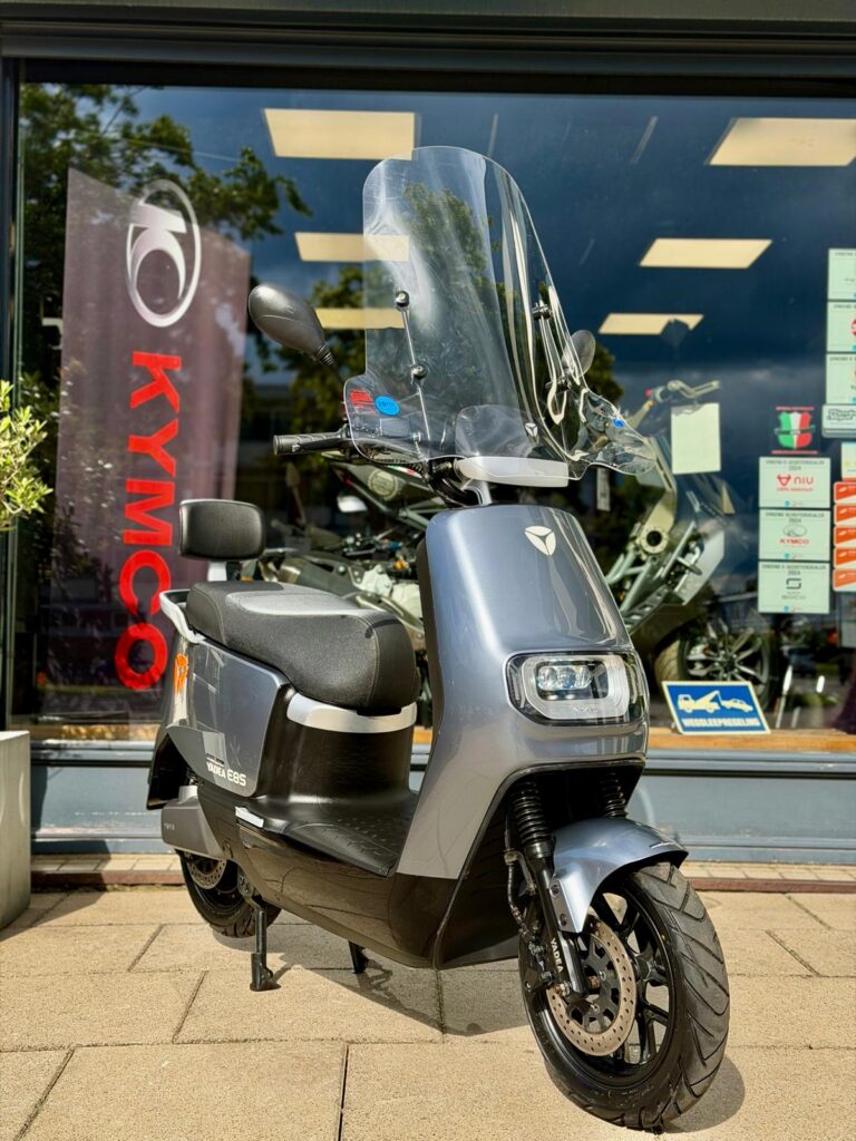 Moderne grijze scooter met windscherm, geparkeerd voor een motorzaak met Kymco-banner.
