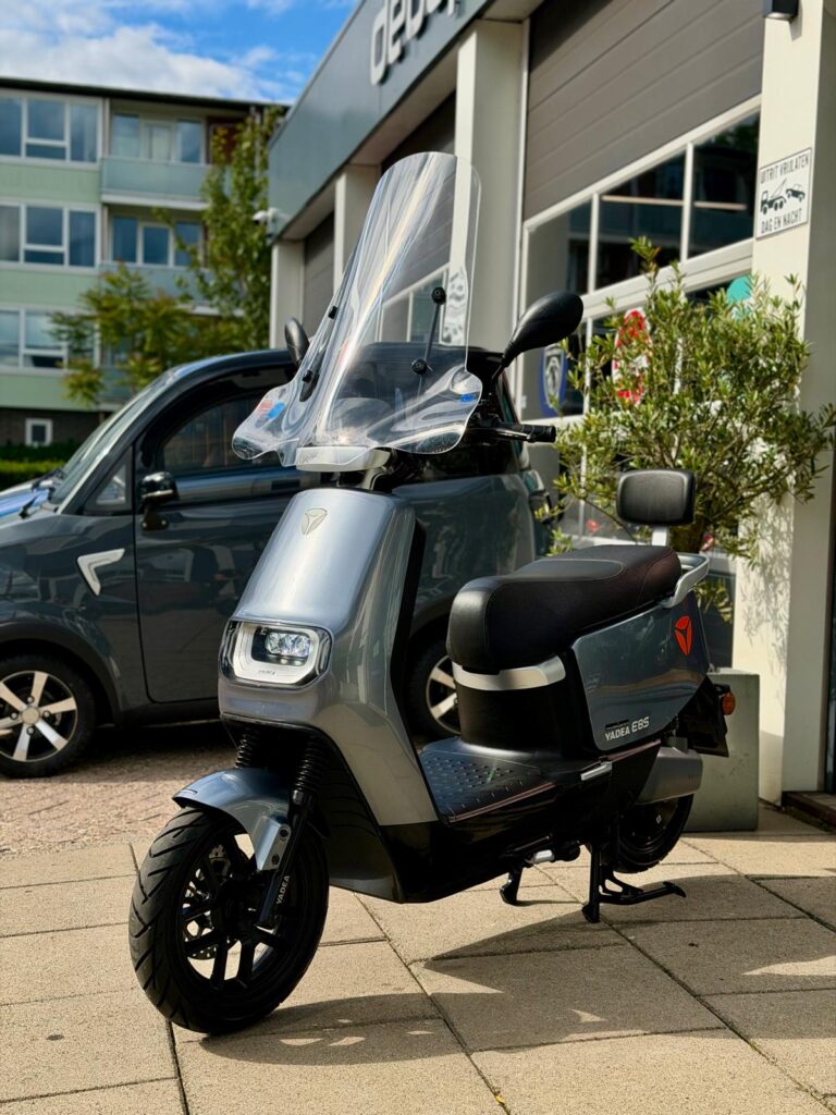 Grijze elektrische scooter met hoog windscherm, Yadea E8S, geparkeerd voor een showroom.