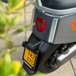 Achterzijde van een grijze scooter met geel NL-kenteken, rode achterlichten en een plant op de voorgrond.