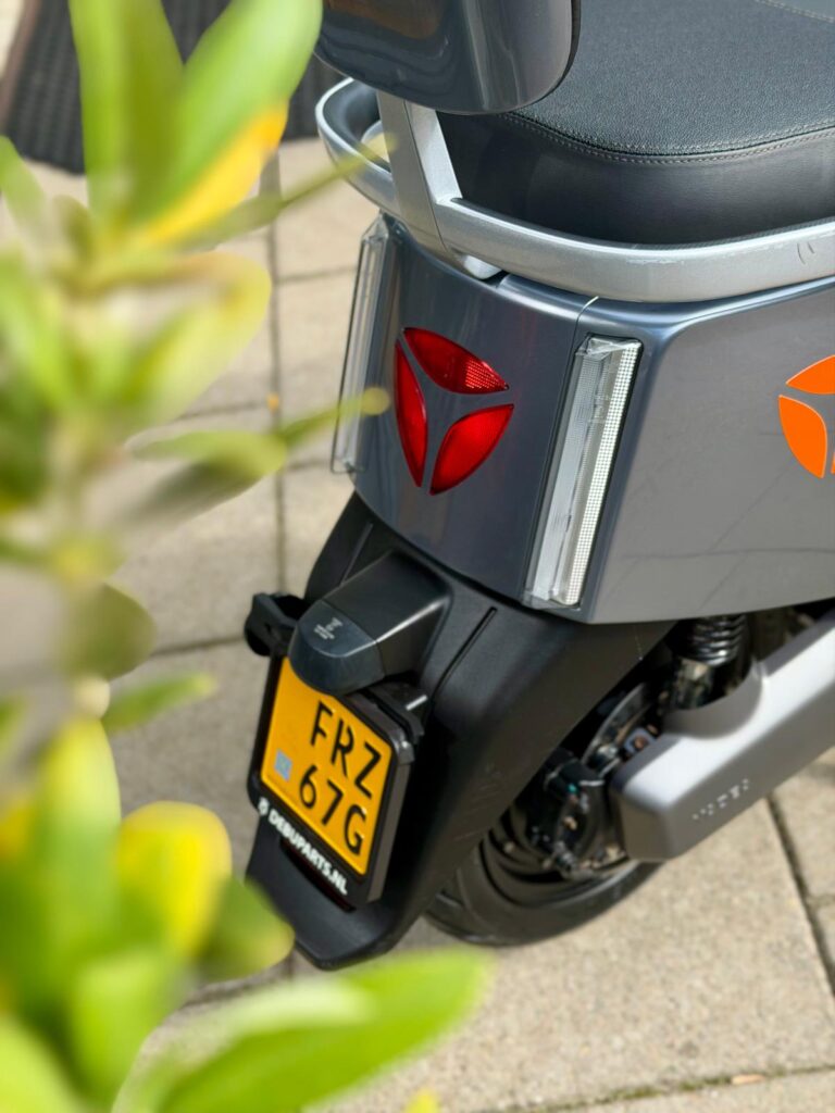 Achterzijde van een grijze scooter met geel NL-kenteken, rode achterlichten en een plant op de voorgrond.