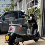 Grijze elektrische scooter Yadea E8S met windscherm, geparkeerd voor een werkplaats.
