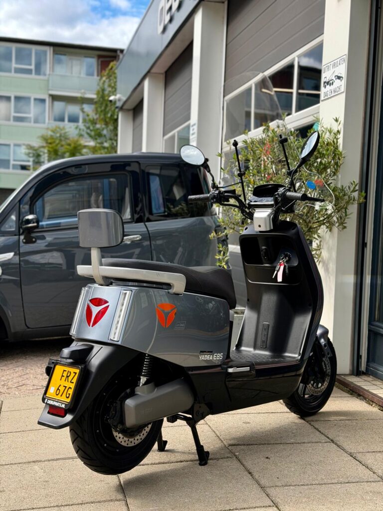 Grijze elektrische scooter Yadea E8S met windscherm, geparkeerd voor een werkplaats.