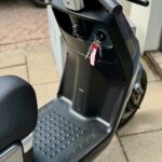 Close-up van een zwarte scooter met sleutels in het contact en antislip voetenplaat, geparkeerd op stoep.