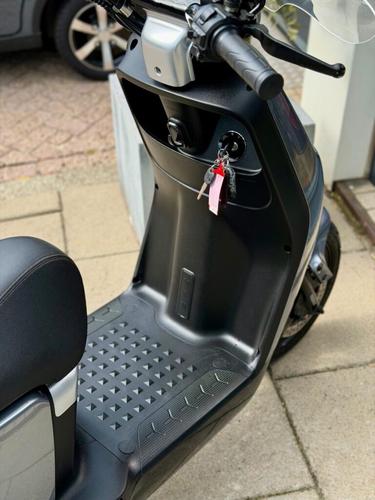Close-up van een zwarte scooter met sleutels in het contact en antislip voetenplaat, geparkeerd op stoep.