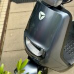Close-up van een grijze elektrische scooter met windscherm, geparkeerd op stoep naast planten.