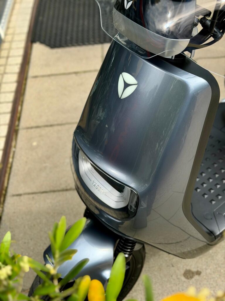Close-up van een grijze elektrische scooter met windscherm, geparkeerd op stoep naast planten.