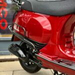 Close-up van rode retro scooter met zwart zadel, uitlaat en bagagerek, op standaard.