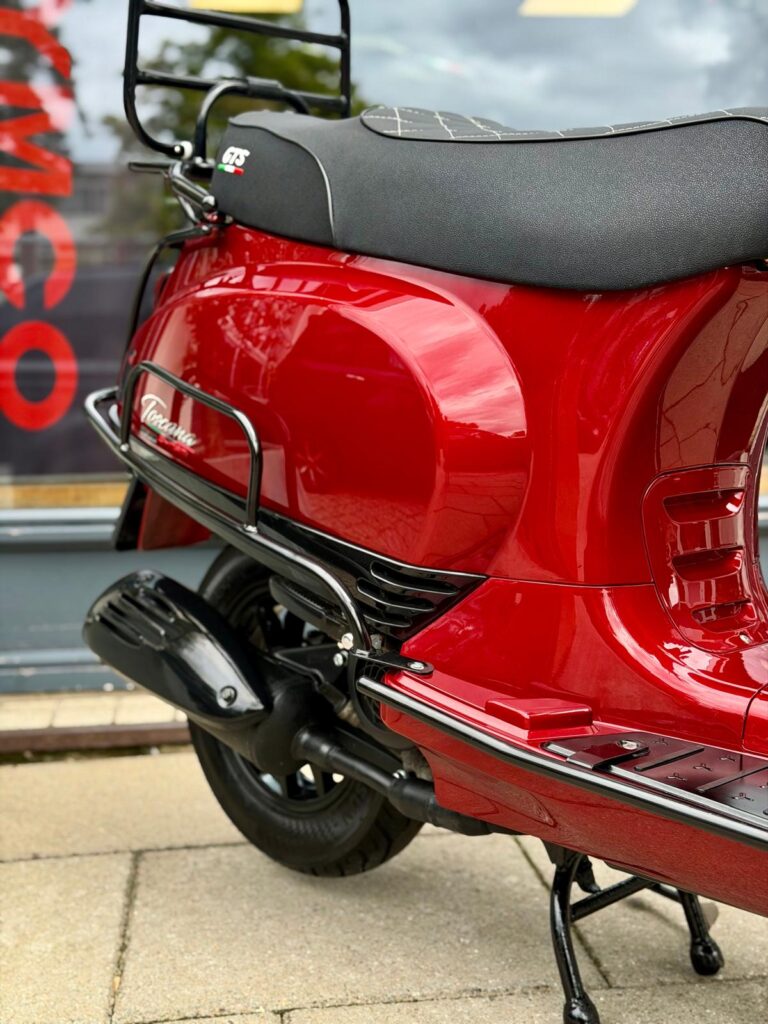 Close-up van rode retro scooter met zwart zadel, uitlaat en bagagerek, op standaard.