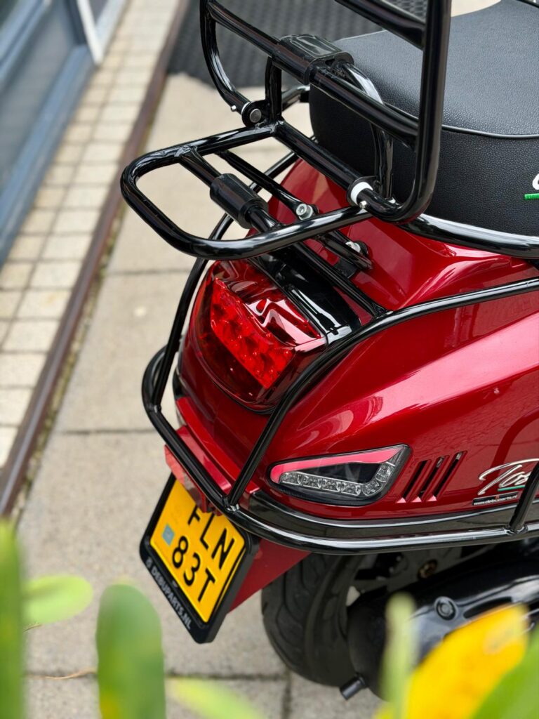 Achterkant van een rode scooter met zwarte bagagedrager, achterlicht en geel NL-kenteken op de stoep.
