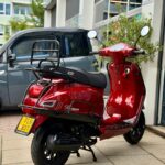 Rode retro scooter met bagagedrager en geel kenteken, geparkeerd voor een pand.