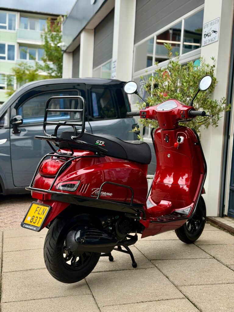 Rode retro scooter met bagagedrager en geel kenteken, geparkeerd voor een pand.