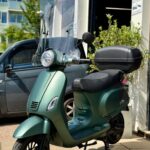 Groene scooter met hoog windscherm en topkoffer, geparkeerd op stoep voor winkel op zonnige dag.
