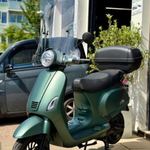 Groene scooter met hoog windscherm en topkoffer, geparkeerd op stoep voor winkel op zonnige dag.