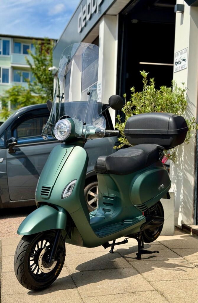 Groene scooter met hoog windscherm en topkoffer, geparkeerd op stoep voor winkel op zonnige dag.