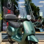 Groene scooter met hoog windscherm en topkoffer, geparkeerd voor een winkelpui.