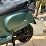Close-up van een matte groene scooter met topkoffer, op de middenbok geparkeerd in de zon.