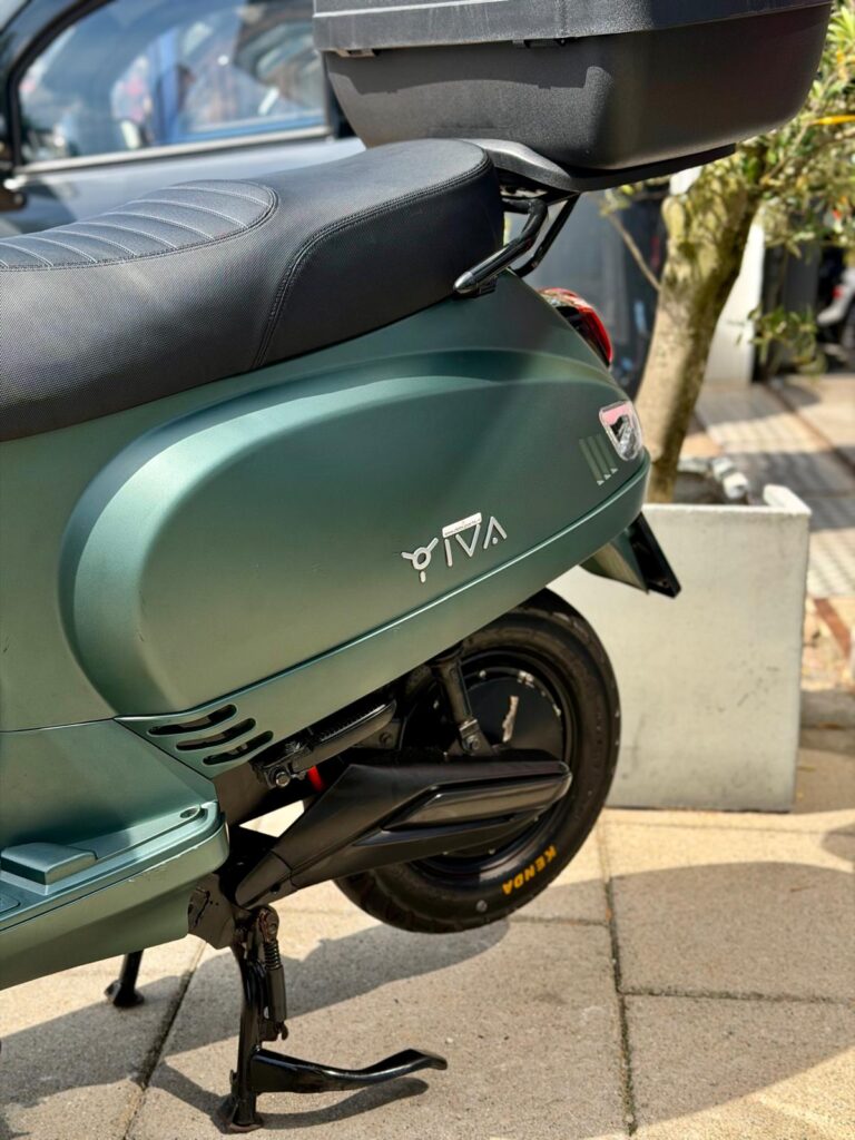 Close-up van een matte groene scooter met topkoffer, op de middenbok geparkeerd in de zon.