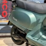 Close-up van een matte groene IVA-scooter met topkoffer, zadel en achterwiel op de standaard voor een winkel.