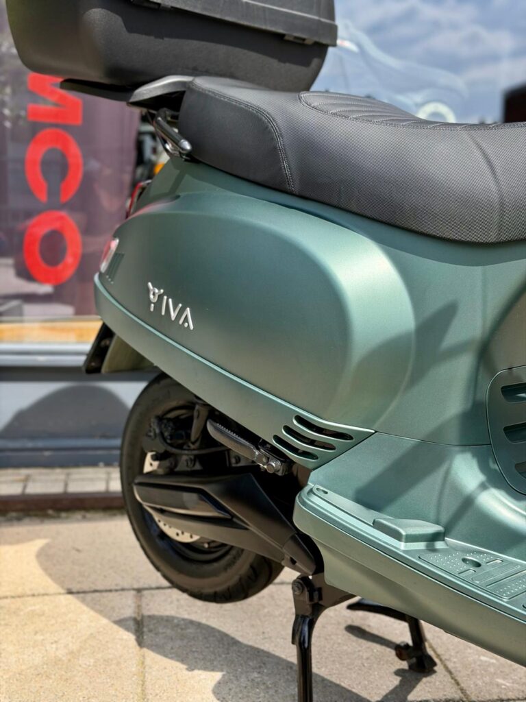 Close-up van een matte groene IVA-scooter met topkoffer, zadel en achterwiel op de standaard voor een winkel.