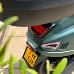 Achterzijde van een matte groene scooter met topkoffer en geel kenteken, tussen planten op de stoep.