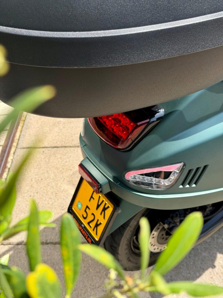 Achterzijde van een matte groene scooter met topkoffer en geel kenteken, tussen planten op de stoep.