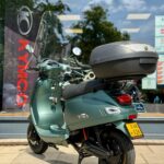 Groene scooter met topkoffer en windscherm geparkeerd voor een scooterwinkel.