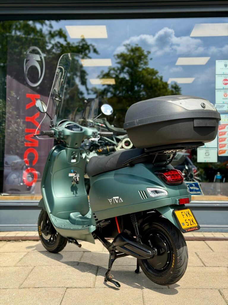 Groene scooter met topkoffer en windscherm geparkeerd voor een scooterwinkel.