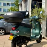 Groene IVA-scooter met topkoffer en windscherm, geparkeerd bij een garage.