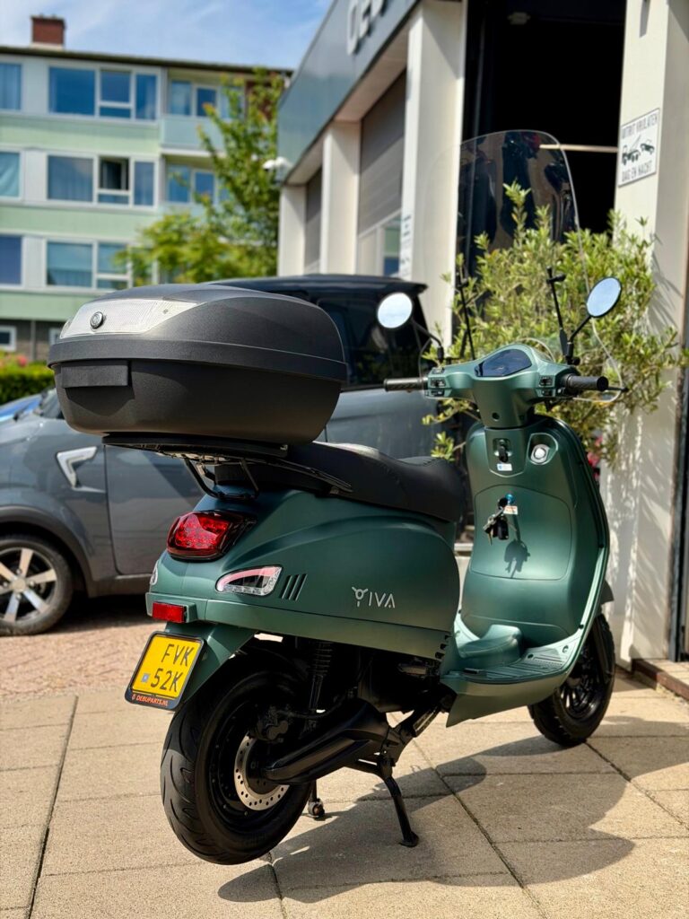 Groene IVA-scooter met topkoffer en windscherm, geparkeerd bij een garage.