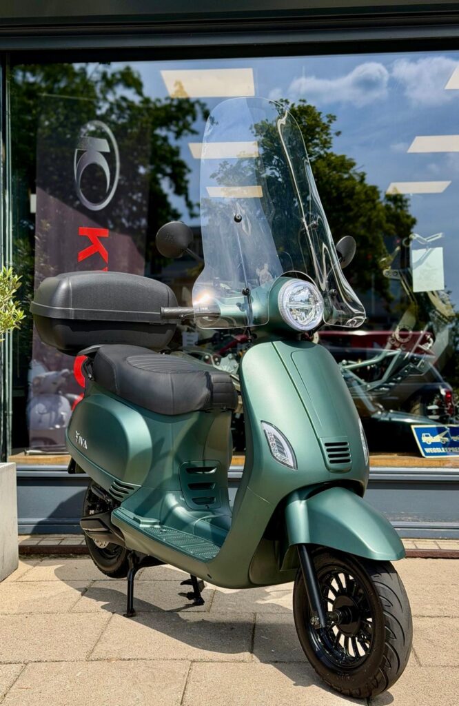 Groene scooter met hoog windscherm en topkoffer, geparkeerd voor een winkelpui.