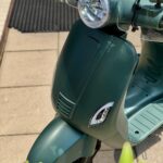 Close-up van een matte groene scooter met ronde koplamp en windscherm op zonnig terras