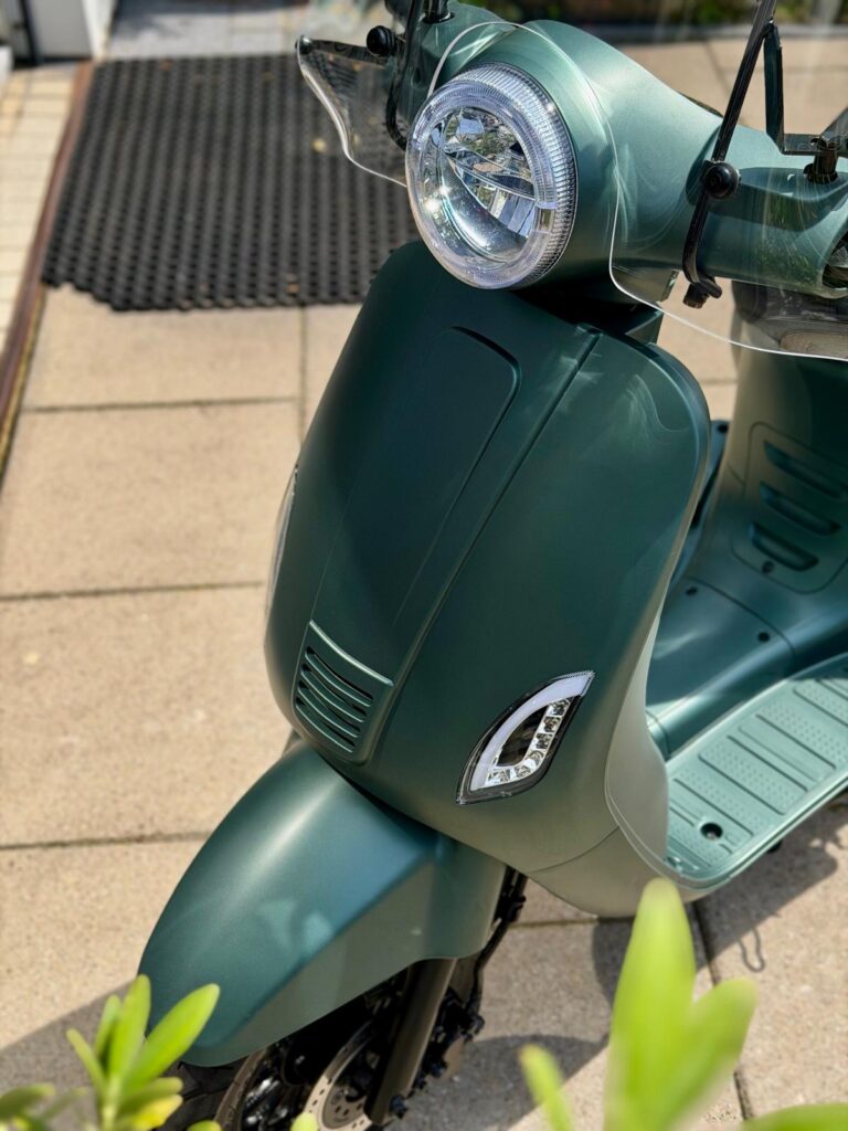 Close-up van een matte groene scooter met ronde koplamp en windscherm op zonnig terras