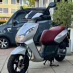 Zilveren scooter met bordeauxrode zitting op standaard, geparkeerd op een stoep bij een gebouw en auto.