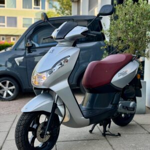 Zilveren scooter met bordeauxrode zitting op standaard, geparkeerd op een stoep bij een gebouw en auto.