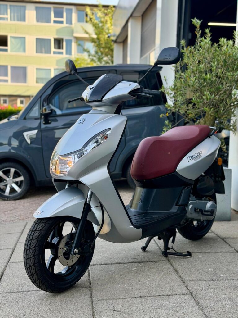 Zilveren scooter met bordeauxrode zitting op standaard, geparkeerd op een stoep bij een gebouw en auto.