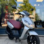 Zilveren scooter met bordeauxrood zadel, geparkeerd voor een winkelruit met weerspiegelde bomen.