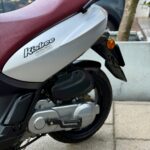 Close-up van de zijkant van een grijze scooter met bordeauxrood zadel, zichtbaar achterwiel en motorblok.