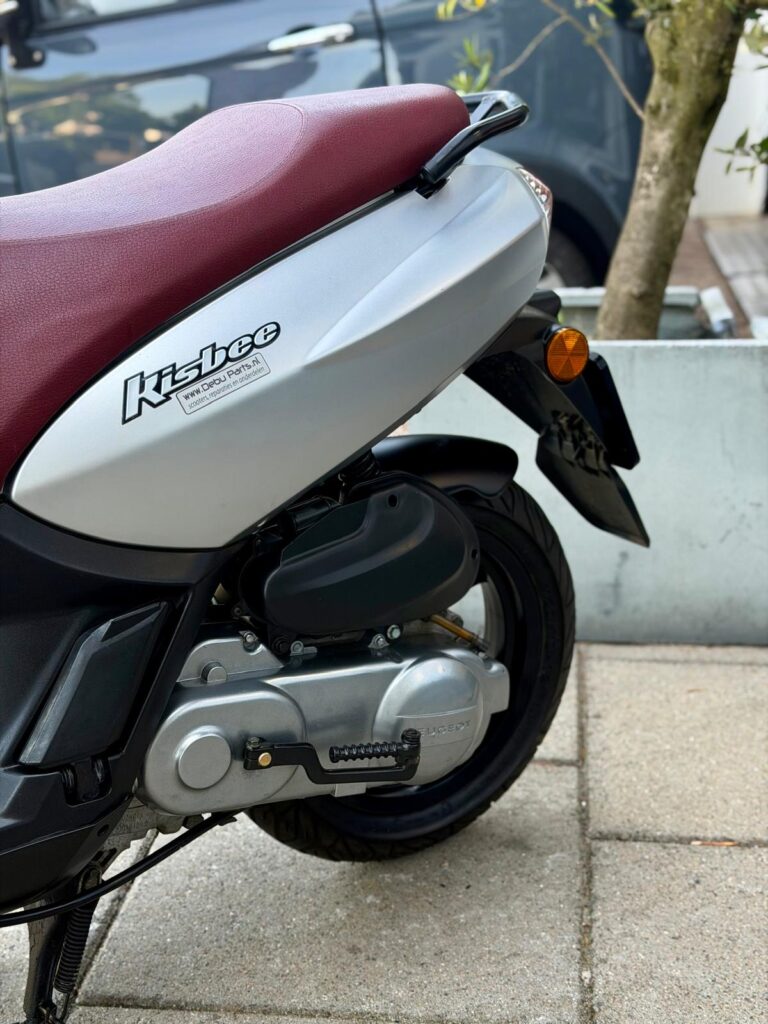 Close-up van de zijkant van een grijze scooter met bordeauxrood zadel, zichtbaar achterwiel en motorblok.