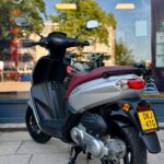 Zilveren scooter met rood zadel geparkeerd voor een winkelraam op een zonnige dag.